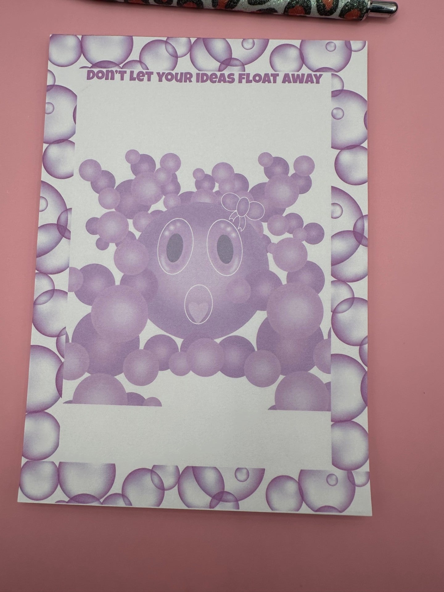 A notepad with bubble character that is purple with a white bow on head and a heart shape mouth surrounded by other size bubbles. The notepad is outlined with purple bubbles and has a heading of "Don't let your ideas float away". The notepad is sitting on a pink background with a pen.