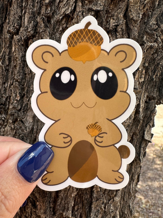 A brown fictional character glossy sticker that has big black eyes and white sparkle. The character is holding an acorn and has an acorn on top of its head. The background has a tree bark setting.