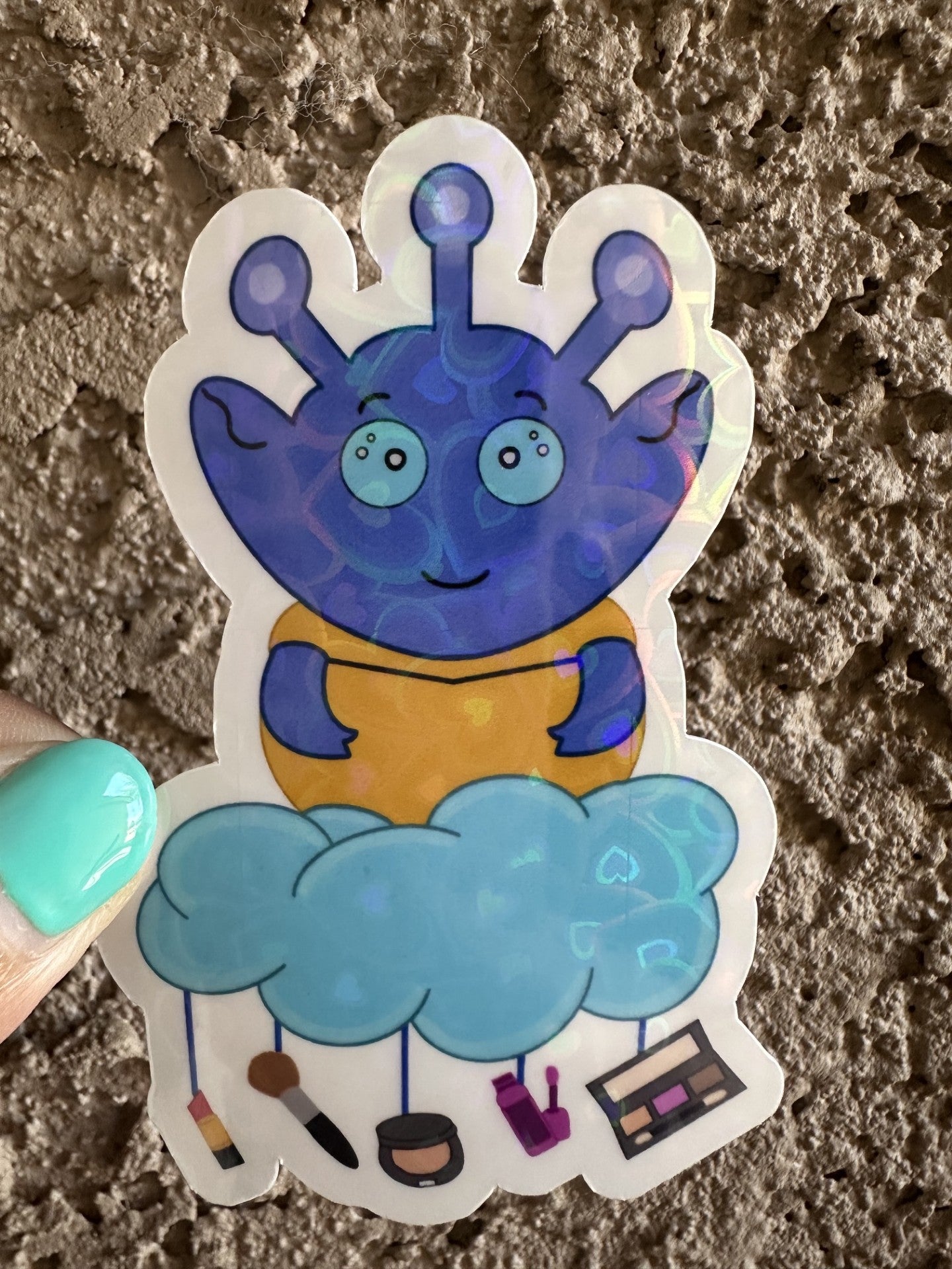 Blue alien sticker that is heart holographic with a cloud and various makeup items hanging from the cloud. The background is a textured surface.