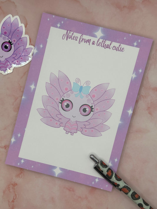 Notepad that has purple and sparkle edges around all four corners. The notepad heading states " Notes from a lethal cutie". The center of the notepad has a fictional character that is purple and pink with wings and a blue bow. The character has big black, purple and pink eyes. The notepad is sitting on top of a pink background with a pen and sticker.