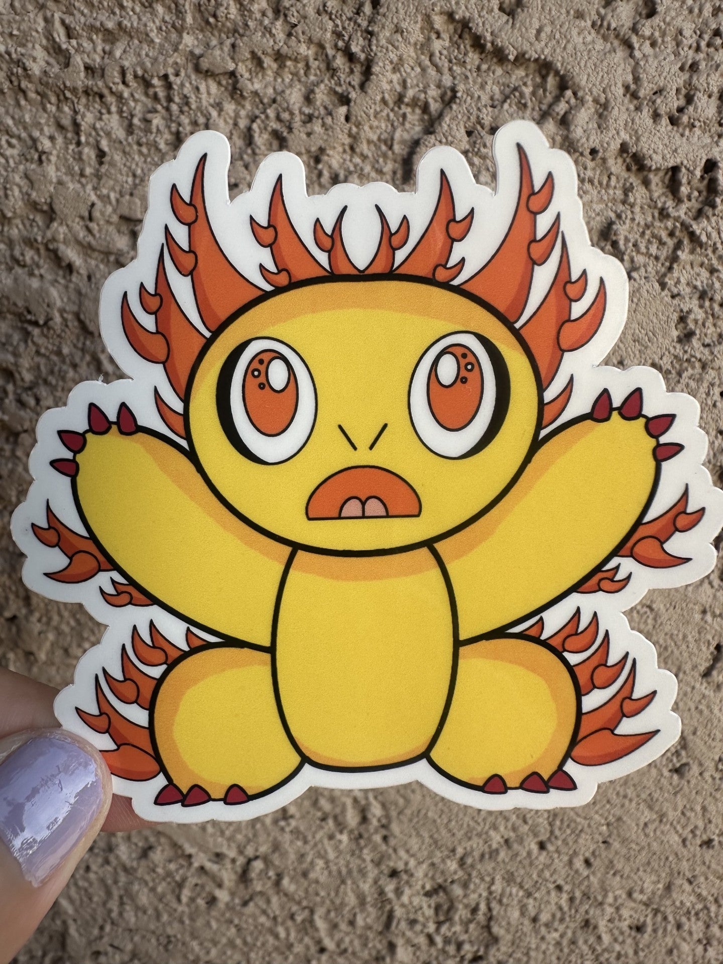Fictional character sticker that is yellow and has multiple spikes coming out of the character that are orange. The character has red nails with big orange eyes with white and black in them.