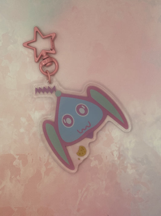 Fictional character keychain that is light blue and turquoise that has big and white eyes, with a purple antenna on head and has a cookie next to it. The keychain has a pink star key ring. The acrylic keychain is sitting on top of a pink textured background.