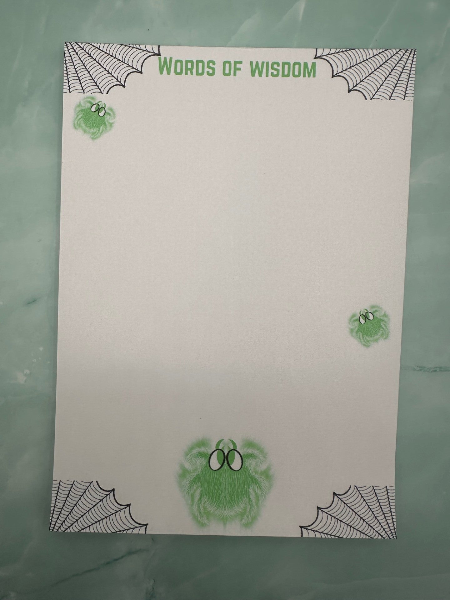 A notepad with green spider character that glows and has white hair with green eyes. Around the four corners of the notepad is spider webs and the heading of the notepad states "Words of wisdom". The notepad is sitting on a light green background.