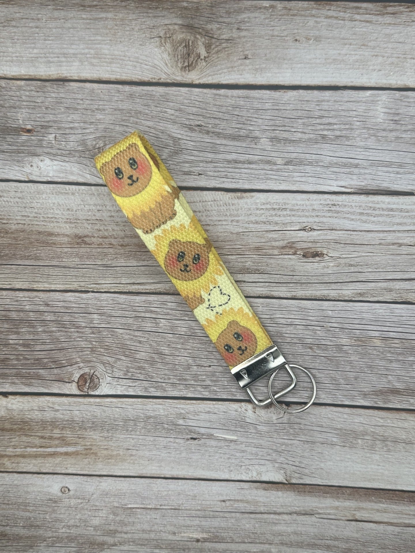 Wristlet keychain with a brown bear that has black eyes and rosy cheeks. The bear has a sunflower shape around its head, and a heart on the bottom of the wristlet. The keychain is on a wooden surface.