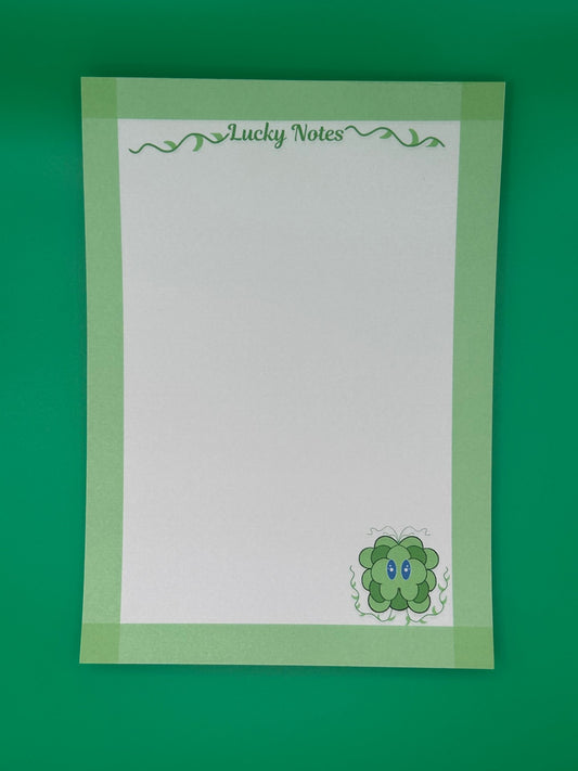 A notepad that has a green leave character at the bottom of the notepad. The character has blue eyes and green vines coming from it. The notepad on all four edges is green and has a heading with vines that states "Lucky notes". The notepad is sitting on top of a green background.