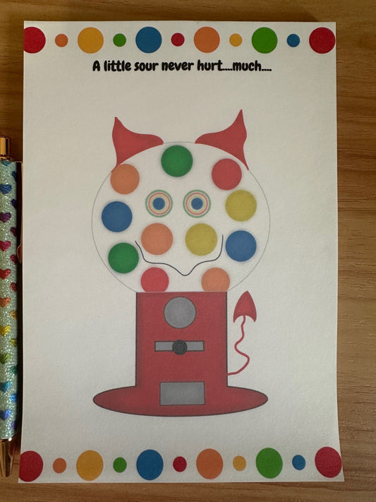 Colorful gumball machine illustration with a sneaky smile that has devil horns and a devil tail. The notepad is on a wooden surface.