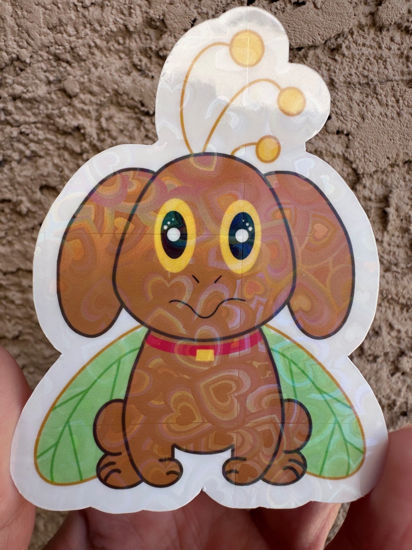 Second option for sticker with heart holographic. Cartoon brown dog that has big yellow and black eyes with green butterfly wings held against a textured beige background.