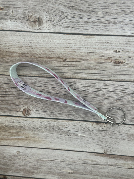 Keychain with a purple cartoon character that is a snake and has dark and light purple colors, with a bow on its head, the wristlet is on a wooden surface side view.
