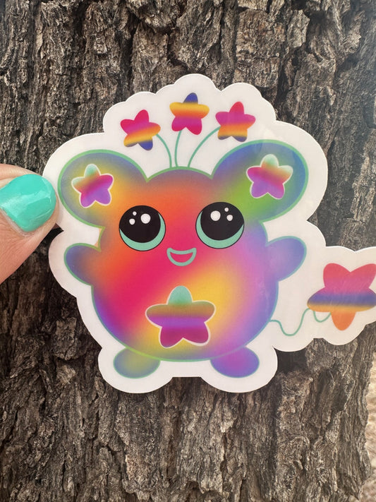 Colorful cartoon character sticker that has stars on the ears and a star on the stomach. The character has different colors like pink, purple, green, and yellow. The character has stars coming from outside of its head and has a star as a tail. The character sticker design is held up against a tree trunk.