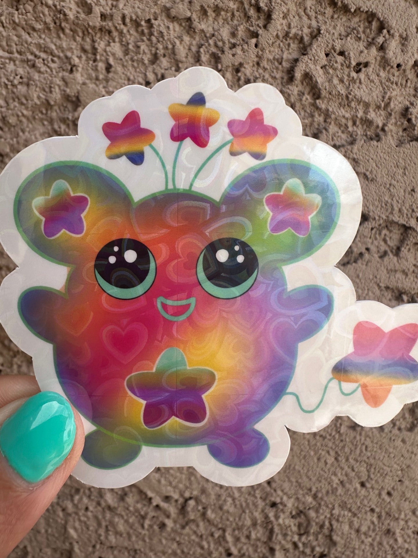 Colorful cartoon character sticker with a heart holographic hat has stars on the ears and a star on the stomach. The character has different colors like pink, purple, green, and yellow. The character has stars coming from outside of its head and has a star as a tail. The character sticker design is held up against a textured surface.