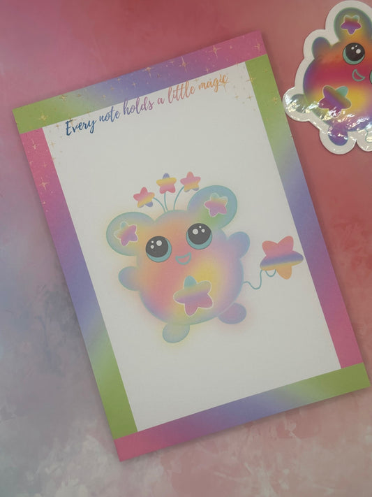 Notepad that has a colorful cartoon character that has stars on the ears and a star on the stomach. The character has different colors like pink, purple, green, and yellow. The character has stars coming from outside of its head and has a star as a tail. The notepad has neon bright colors on all four corners and has sparkling stars on top of the notepad. The notepad has the heading "Every note holds a little magic". The notepad is sitting on a bright background with a sticker.