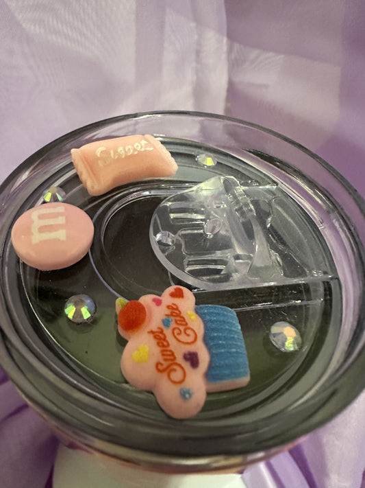A clear snow globe tumbler lid featuring several floating faux candy decorations and iridescent gems. One large pink candy piece has 'Sweet Cake' written in red script, and another rectangular pink piece says 'Sweet'.
