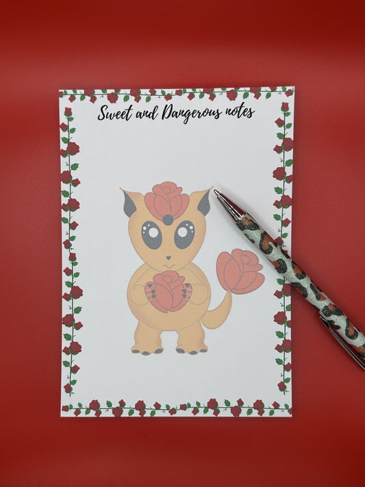 Notepad with a cute fictional creature design that is orange and has a rose on its head and a tail. The orange creature is holding a red rose and has big black eyes and black ears. The notepad has roses all around the four corners of the notepad and has a notepad heading of "Sweet and Dangerous Notes". The notepad is sitting on a red background with a pen.