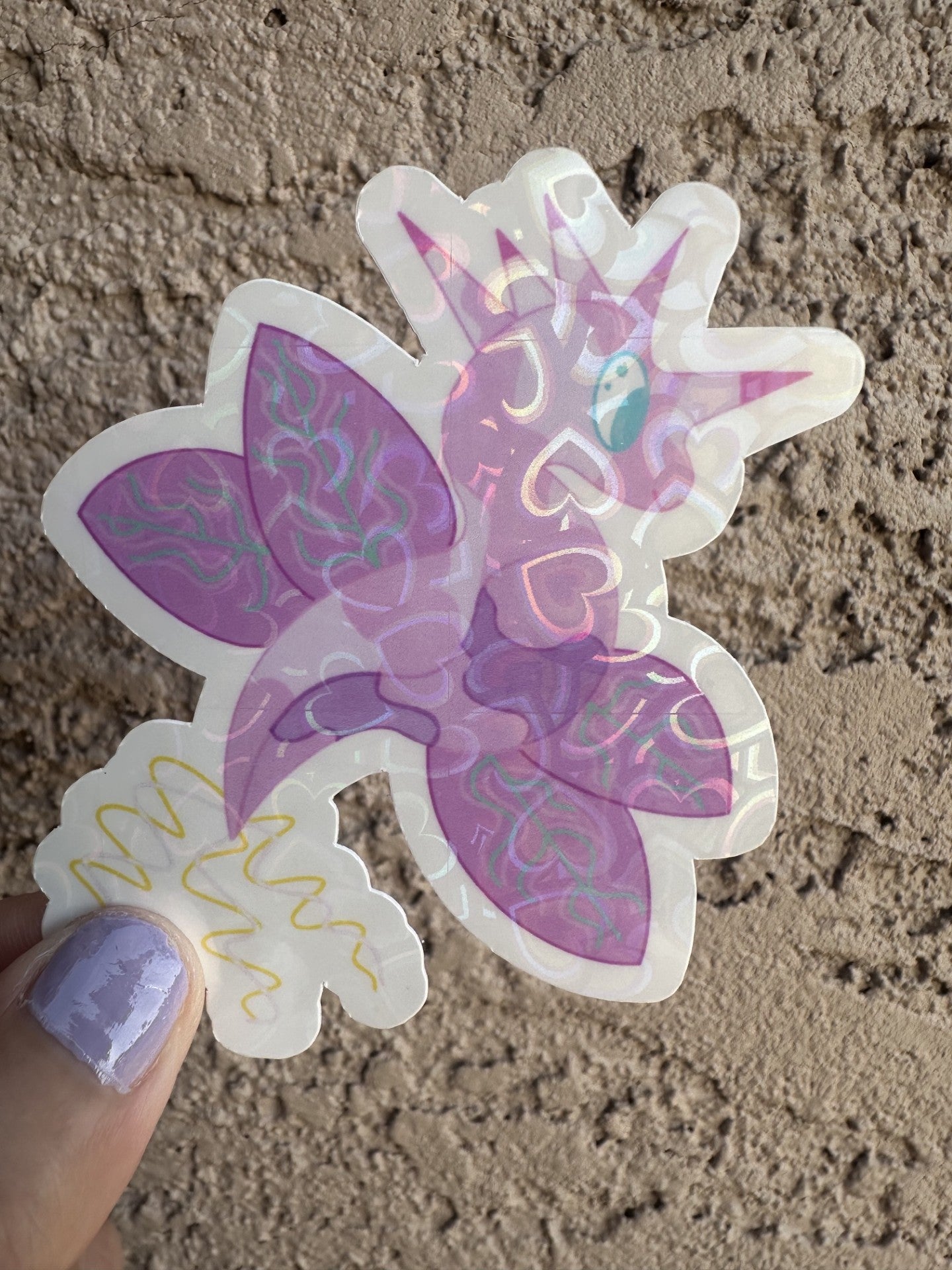 Fictional heart holographic character sticker that is light purple and dark purple, that looks like a dragon and also has wings with turquoise lines. The character has five horns coming from its head that are pink and purple, while the tail is yellow and light pink. The character has light blue eyes.
