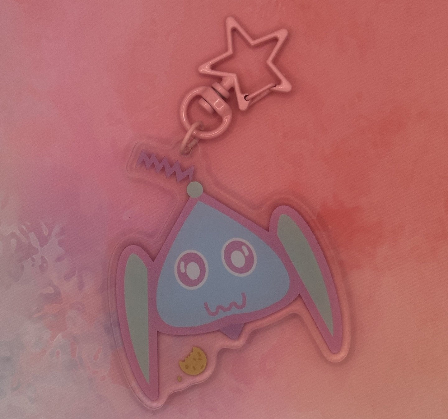 Fictional character keychain that is light blue and turquoise that has big and white eyes, with a purple antenna on head and has a cookie next to it. The keychain has a pink star key ring. The acrylic keychain is sitting on top of a pink textured background.