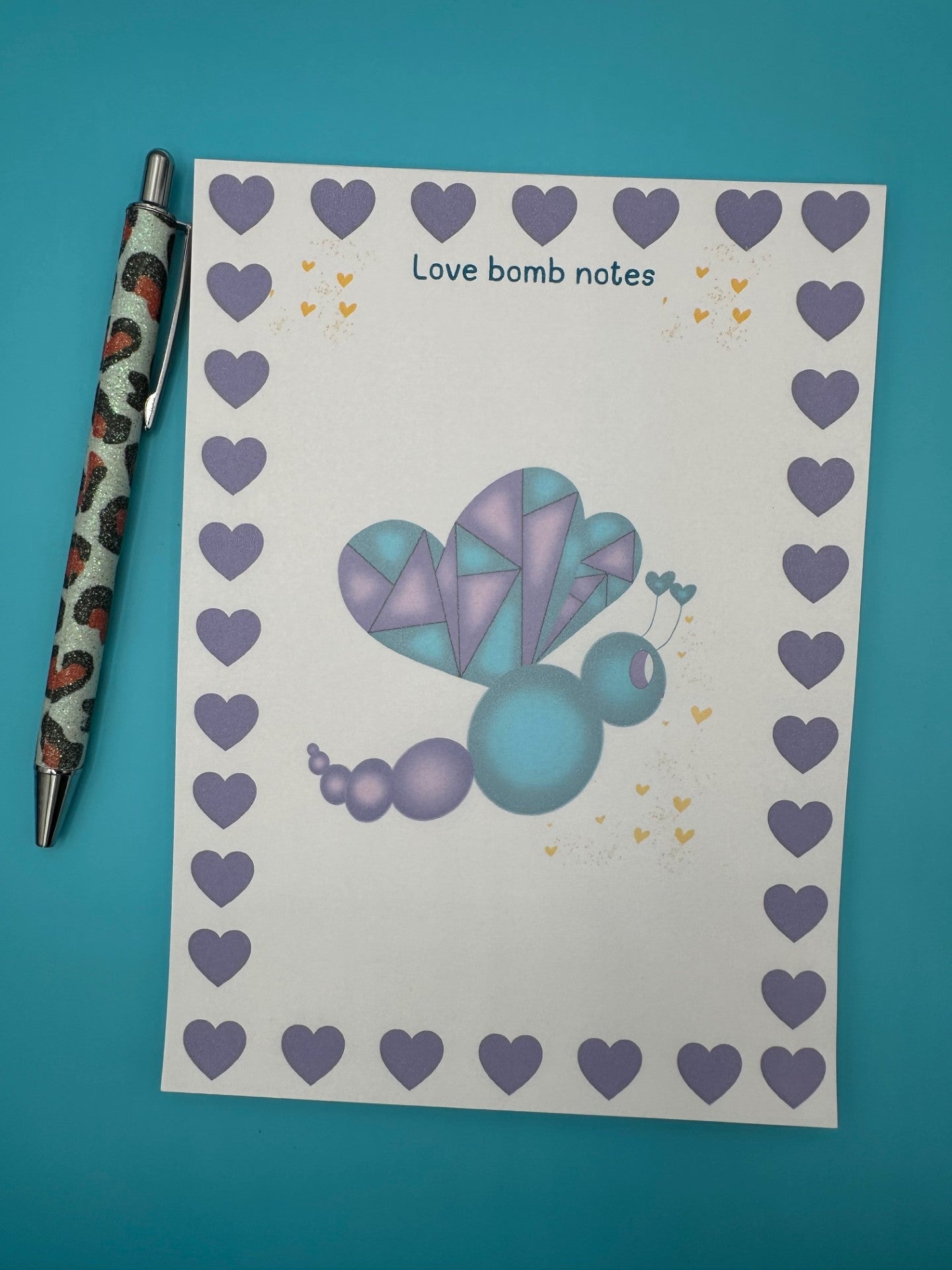A notepad with a colorful butterfly illustration with purple and dark blue colors and a heart border. The notepad is on a blue background, accompanied by a pen.