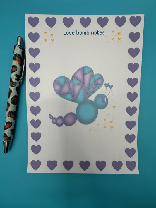 A notepad with a colorful butterfly illustration with purple and dark blue colors and a heart border. The notepad is on a blue background, accompanied by a pen.