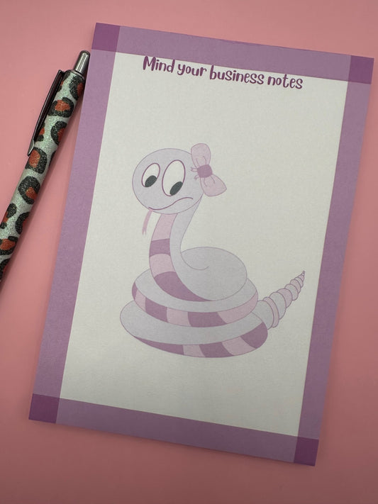 A notepad with fictional character is that a dark and light pink color with a bow on top of the head and the tongue sticking out. The notepad has purple edges and the notepad heading stating "mind your business notes".