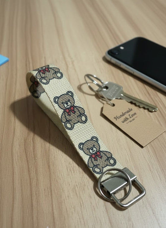 Keychain with teddy bear pattern on a wooden surface next to a phone and key.