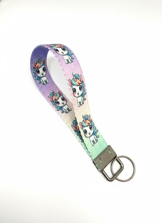 A fabric wristlet keychain with a purple, blue, and green gradient background, patterned with cartoon unicorns and featuring a silver-toned metal key ring attachment.