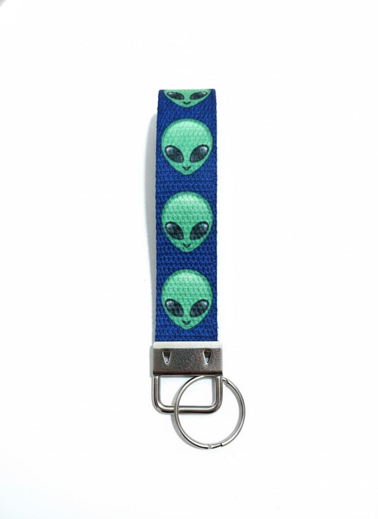 A dark blue fabric wristlet keychain patterned with green alien heads, featuring a silver-toned metal key ring attachment.