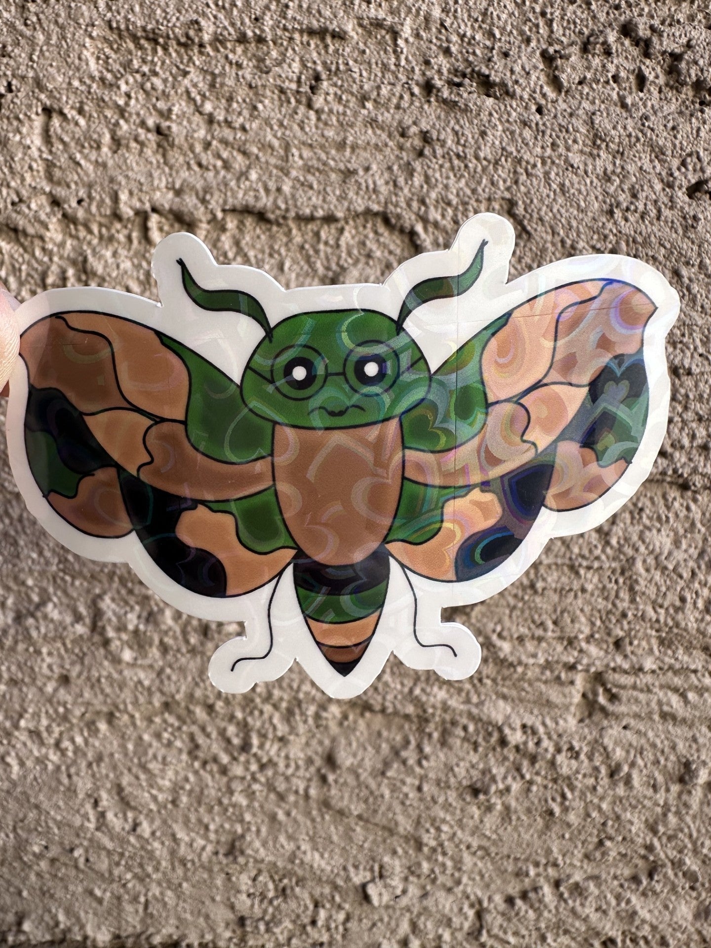 A fictional character sticker that has camouflage color that is an animal wasp shape and has black colored glasses. The sticker has a heart holographic. 