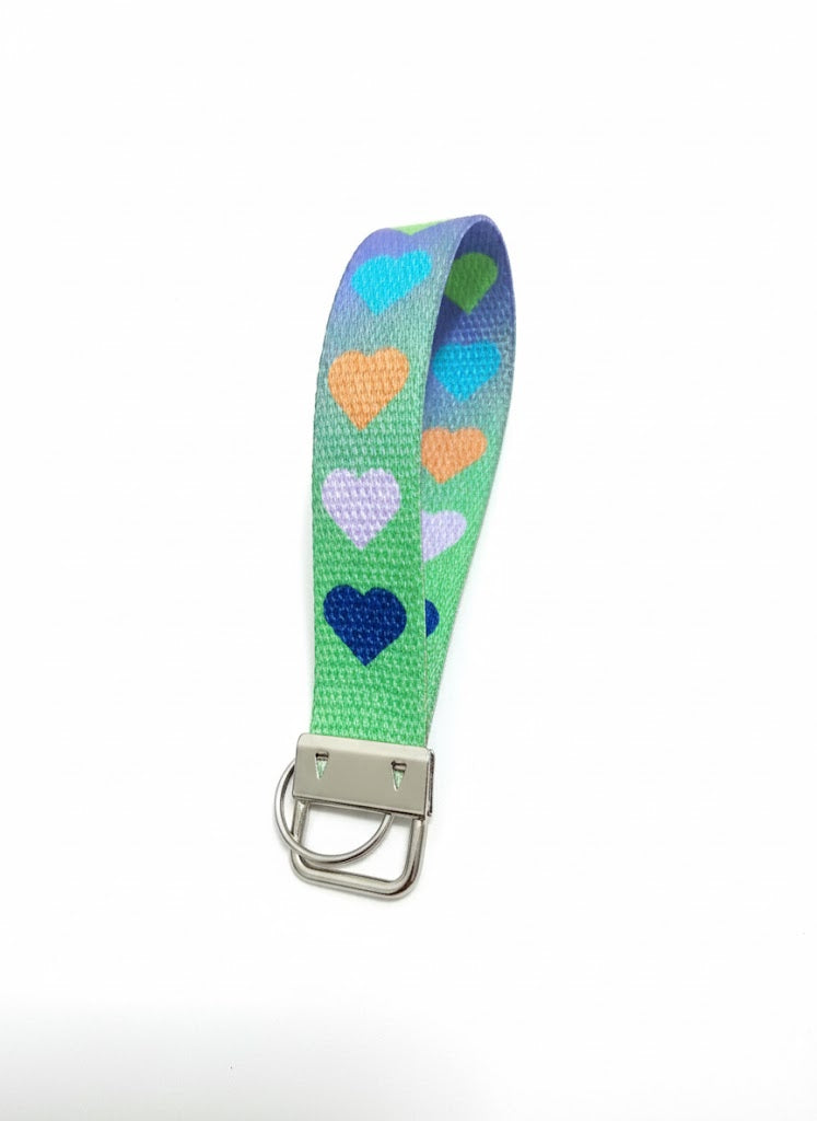 A purple fabric wristlet keychain featuring a vertical pattern of turquoise, orange, white, and dark blue hearts.