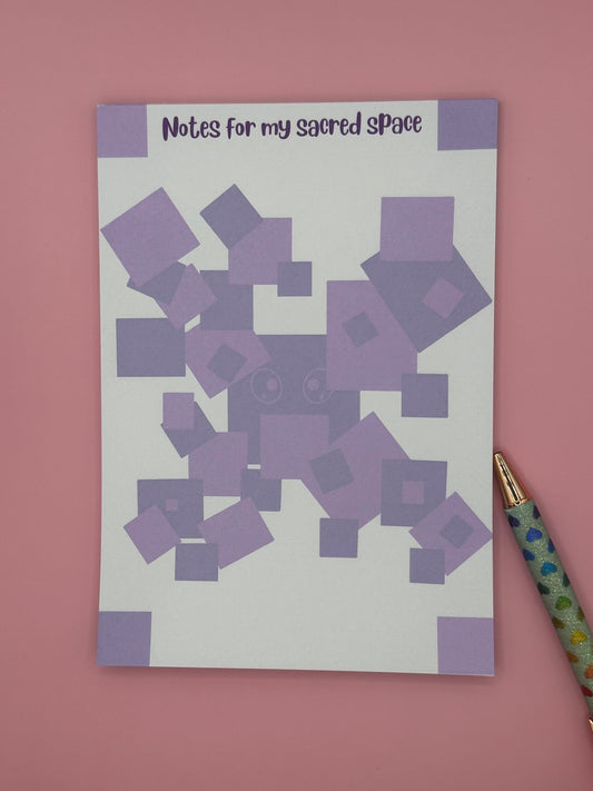 A notepad that has a purple boxes as a character with eyes. The notepad has square edges around the four corners of the notepad and the heading of the notepad stating "Notes for my sacred space". The notepad is sitting on top of a pink background with a pen. 