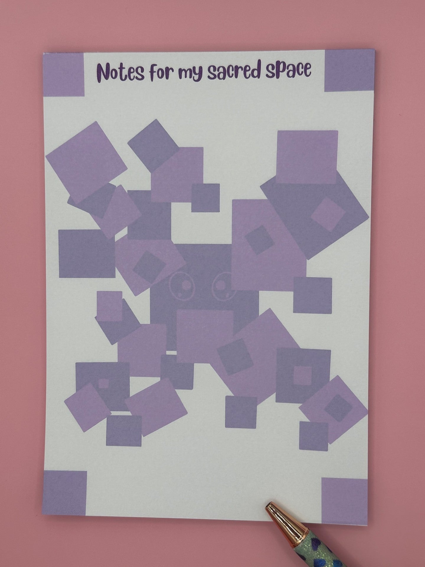 A notepad that has a purple boxes as a character with eyes. The notepad has square edges around the four corners of the notepad and the heading of the notepad stating "Notes for my sacred space". The notepad is sitting on top of a pink background with a pen. 