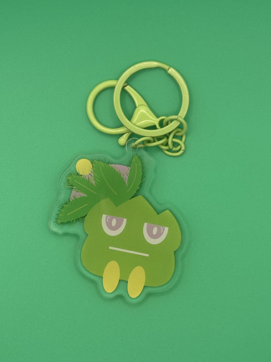Green cartoon character keychain on a green background. The character has green leaves coming from it with yellow and purple on top of the green leaves. The character has a serious face with purple and white eyes. The keychains has a green keyring. 