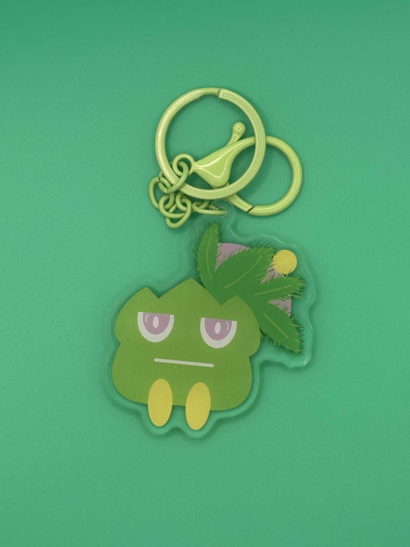 Green cartoon character keychain on a green background. The character has green leaves coming from it with yellow and purple on top of the green leaves. The character has a serious face with purple and white eyes. The keychains has a green keyring. 