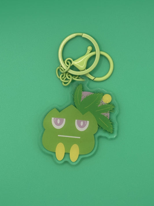 Green cartoon character keychain on a green background. The character has green leaves coming from it with yellow and purple on top of the green leaves. The character has a serious face with purple and white eyes. The keychains has a green keyring. 
