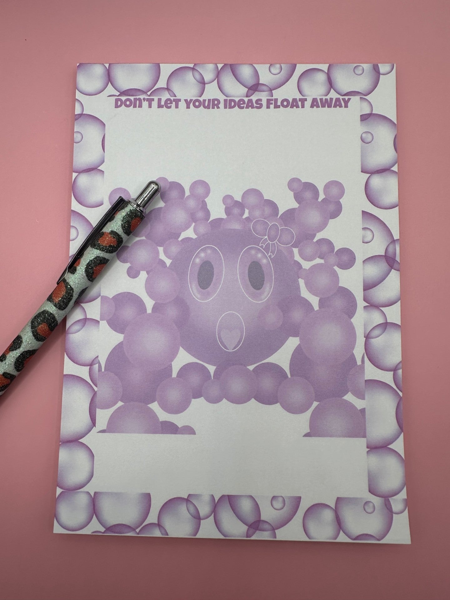 A notepad with bubble character that is purple with a white bow on head and a heart shape mouth surrounded by other size bubbles. The notepad is outlined with purple bubbles and has a heading of "Don't let your ideas float away". The notepad is sitting on a pink background with a pen.