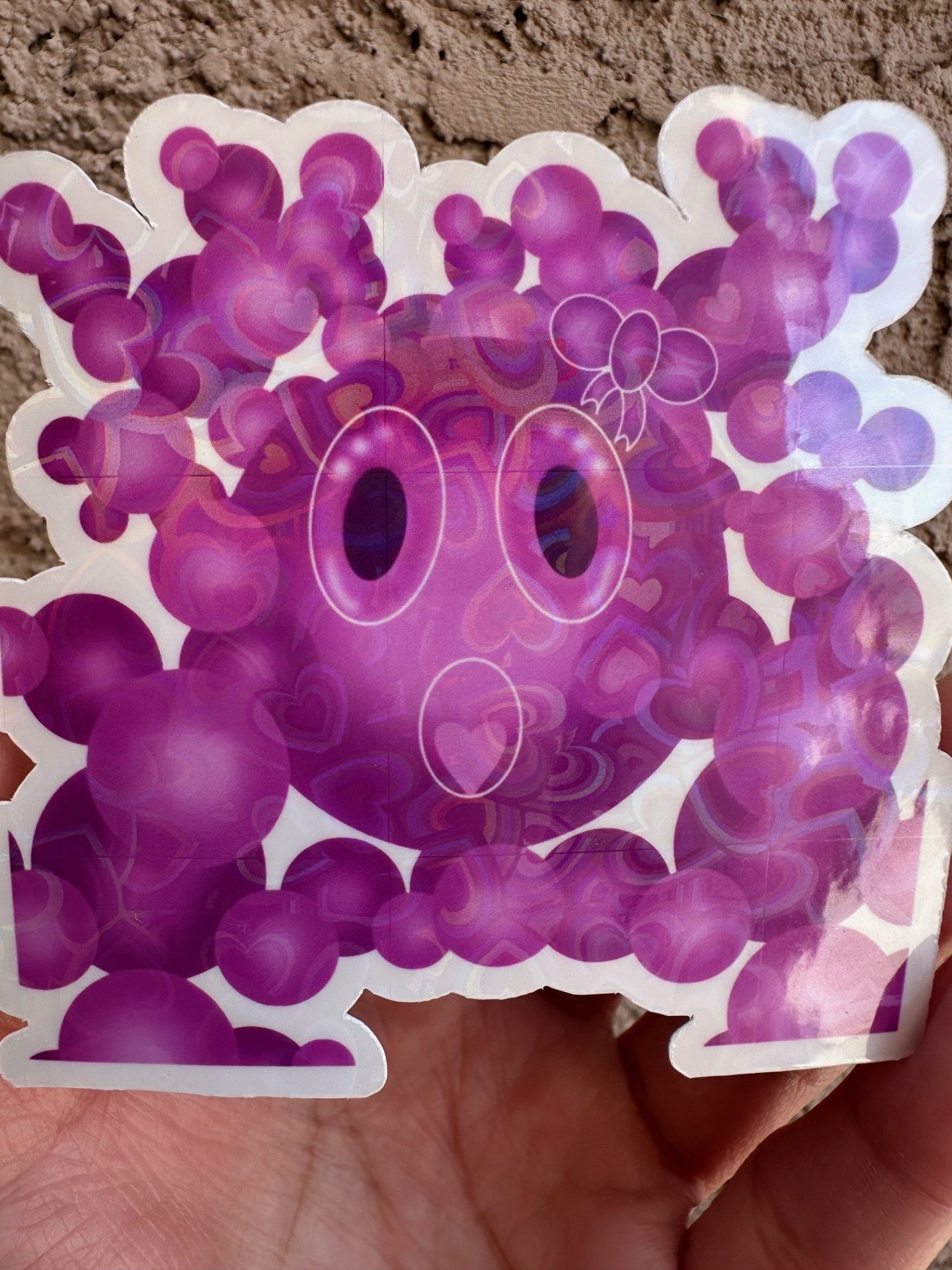 Second option for sticker with heart holographic design. A Purple cartoon character that has a bow on the head with big bubble eyes and a heart mouth surrounded by bubbles held against a wall.