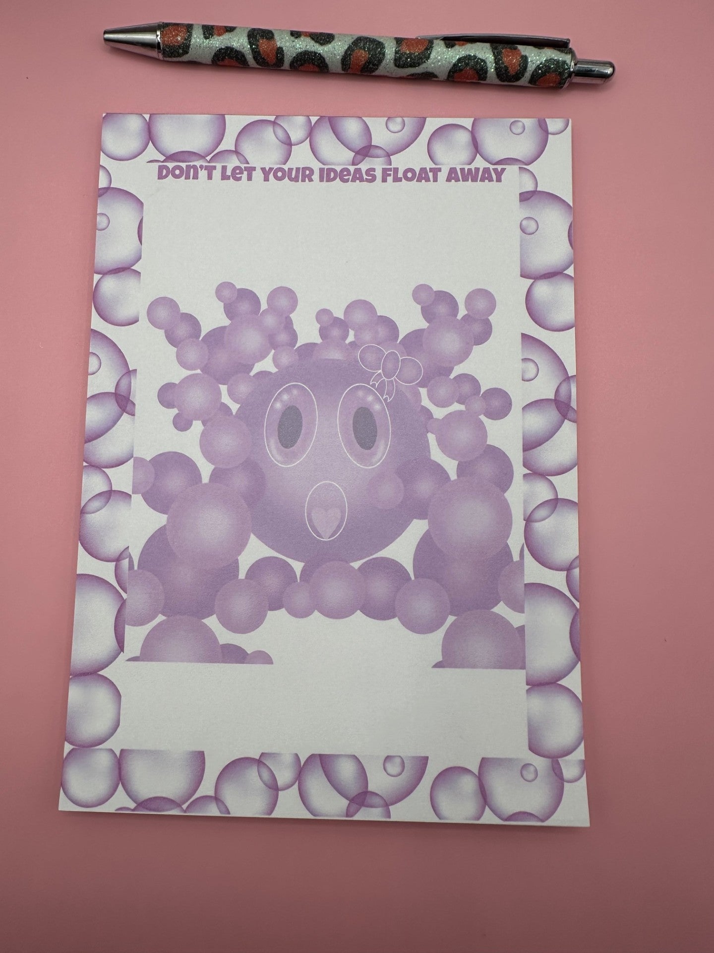 A notepad with bubble character that is purple with a white bow on head and a heart shape mouth surrounded by other size bubbles. The notepad is outlined with purple bubbles and has a heading of "Don't let your ideas float away". The notepad is sitting on a pink background with a pen.