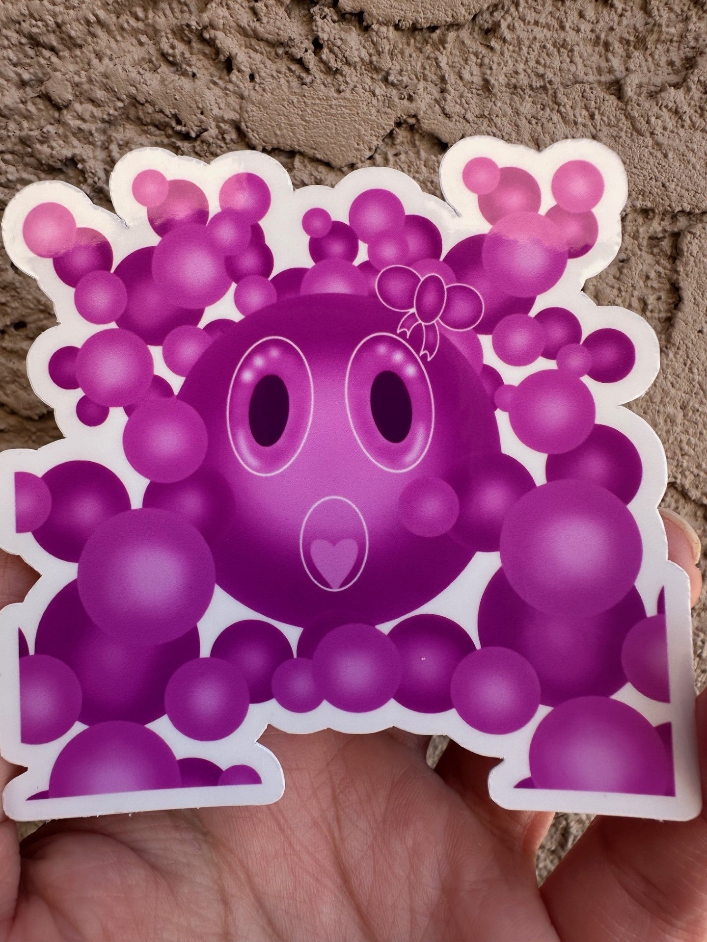Purple cartoon character that has a bow on the head with big bubble eyes and a heart mouth surrounded by bubbles held against a wall.