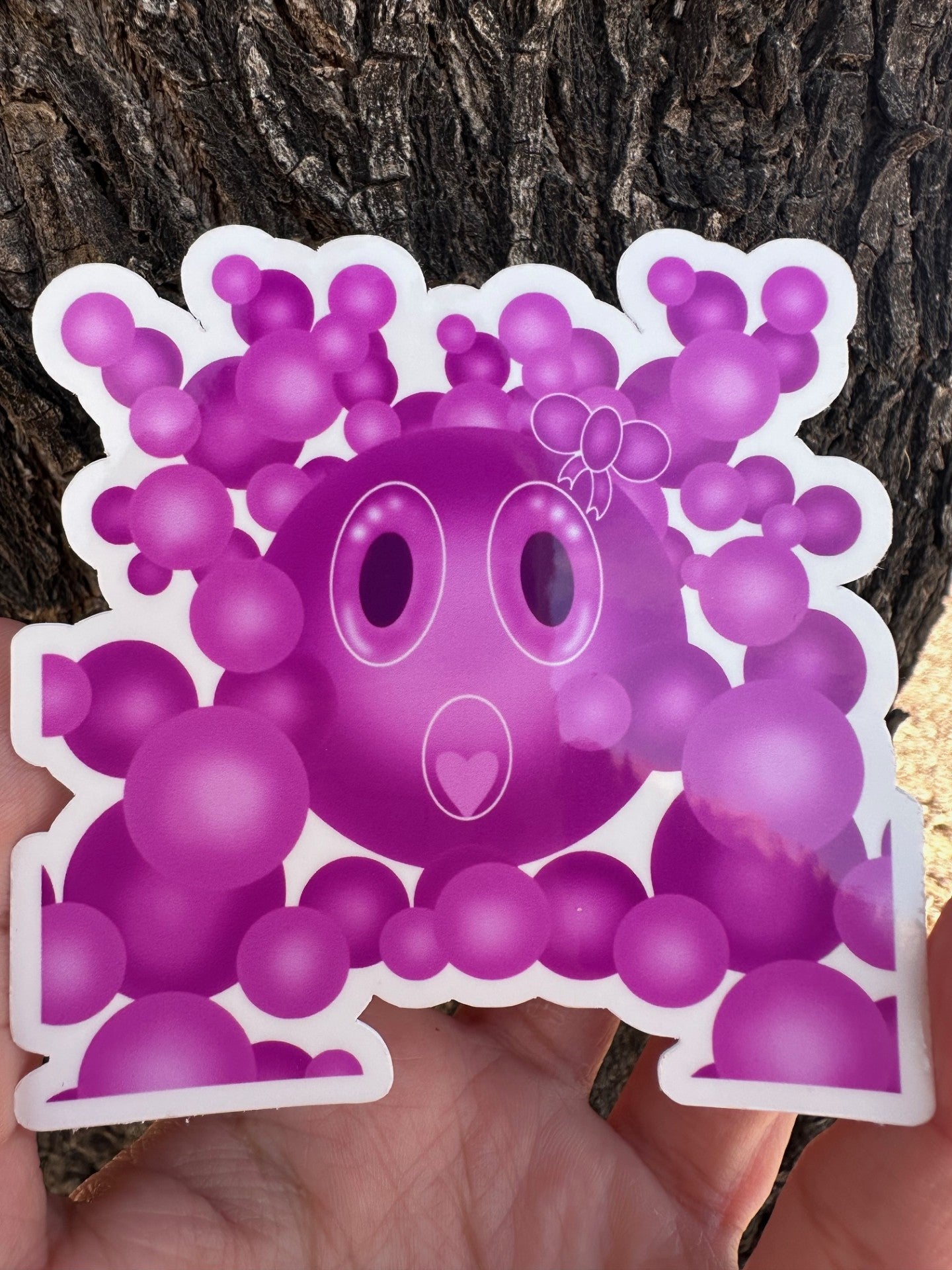 Purple cartoon character that has a bow on the head with big bubble eyes and a heart mouth surrounded by bubbles held against a tree bark background.
