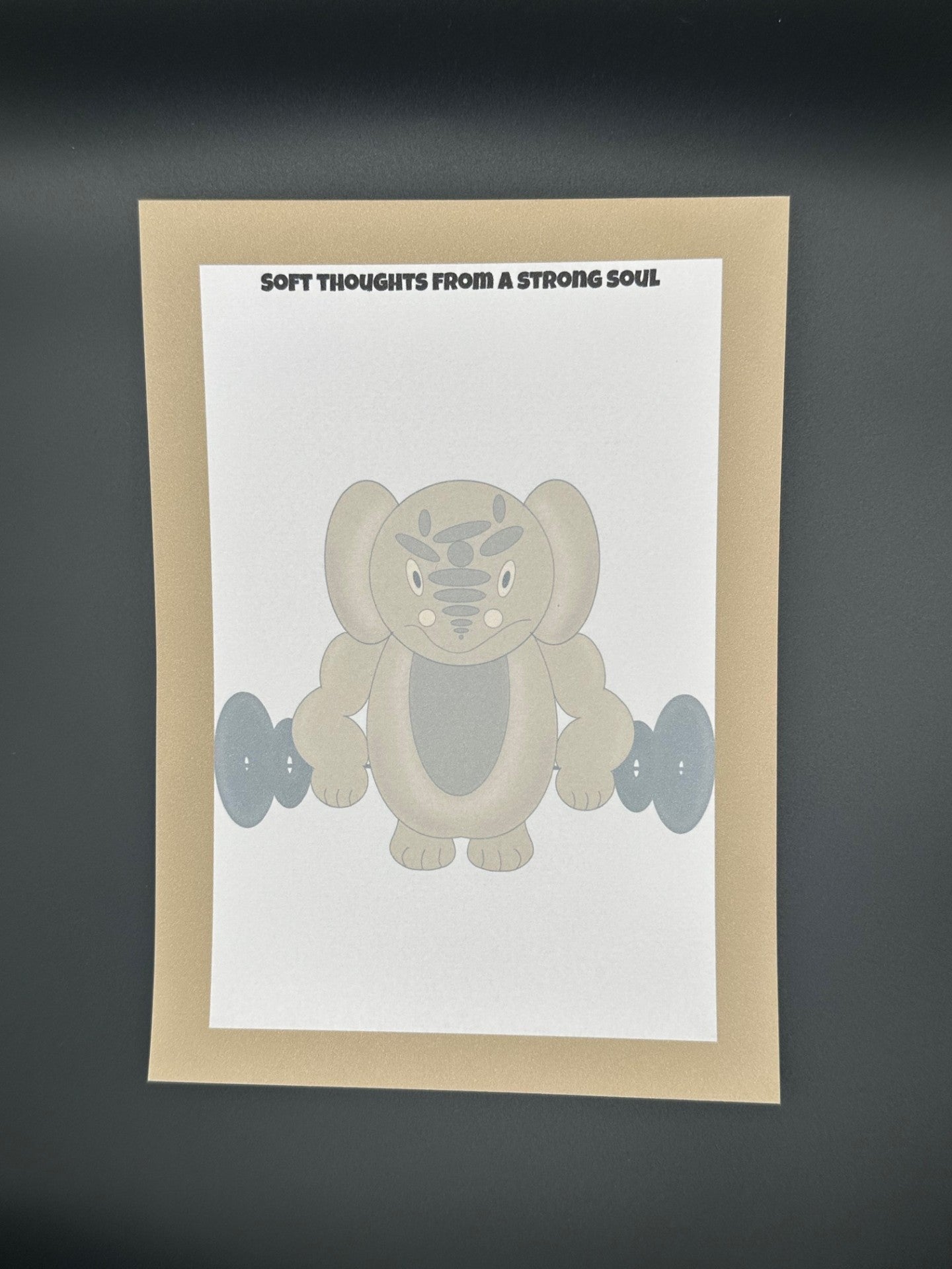 A notepad that has a brown character that is a rounded shape with muscles and has black eyes and a smile. The character is holding black weights. The notepad has brown edges all around the four corners and a notepad heading stating " Soft thoughts from a strong soul." The notepad is sitting on top of a black background.
