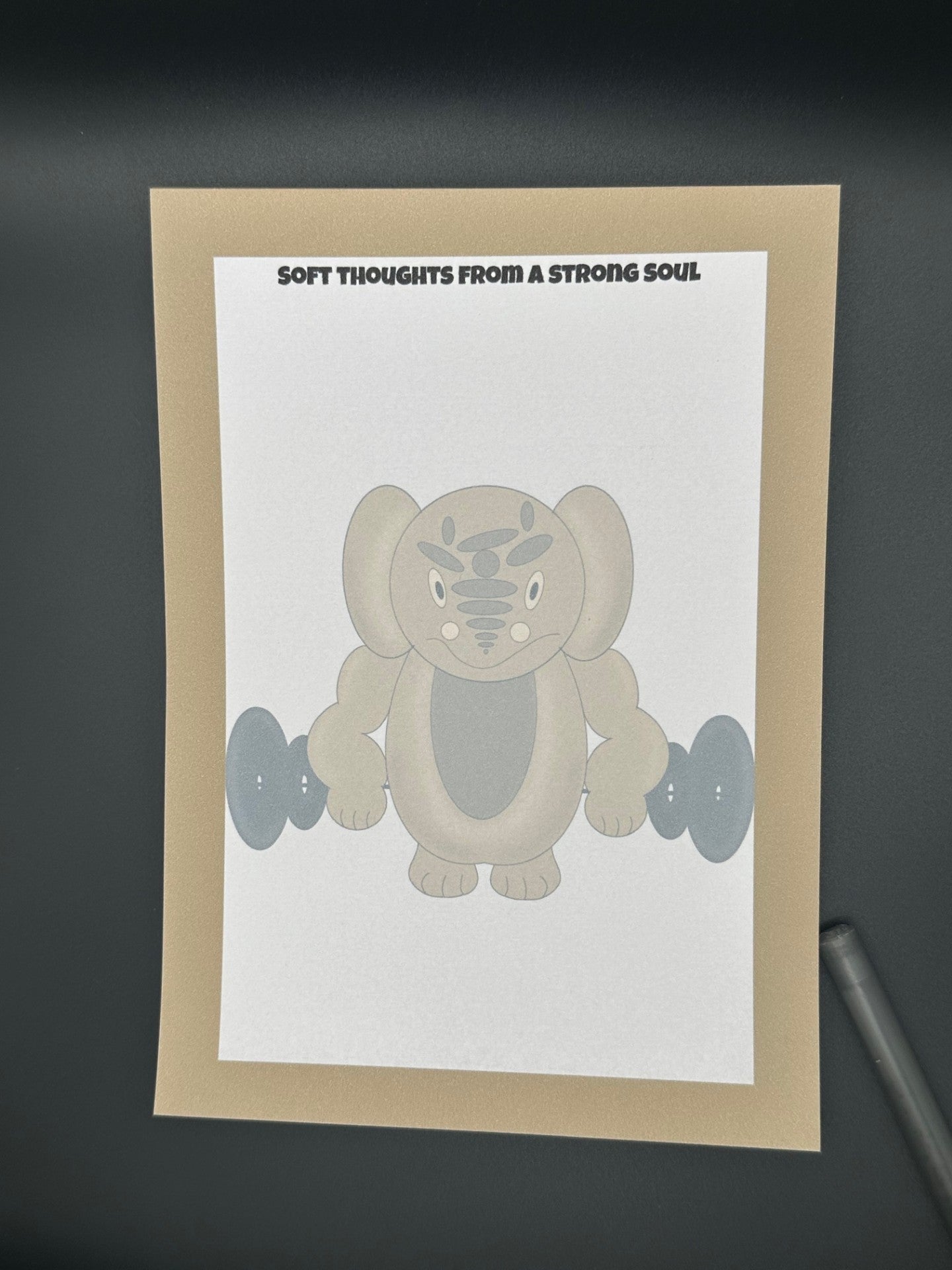 A notepad that has a brown character that is rounded shape with muscles and has black eyes and a smile. The character is holding black weights. The notepad has brown edges all around the four corners and a notepad heading stating " Soft thoughts from a strong soul" The notepad is sitting on top of a black background with a pen. 