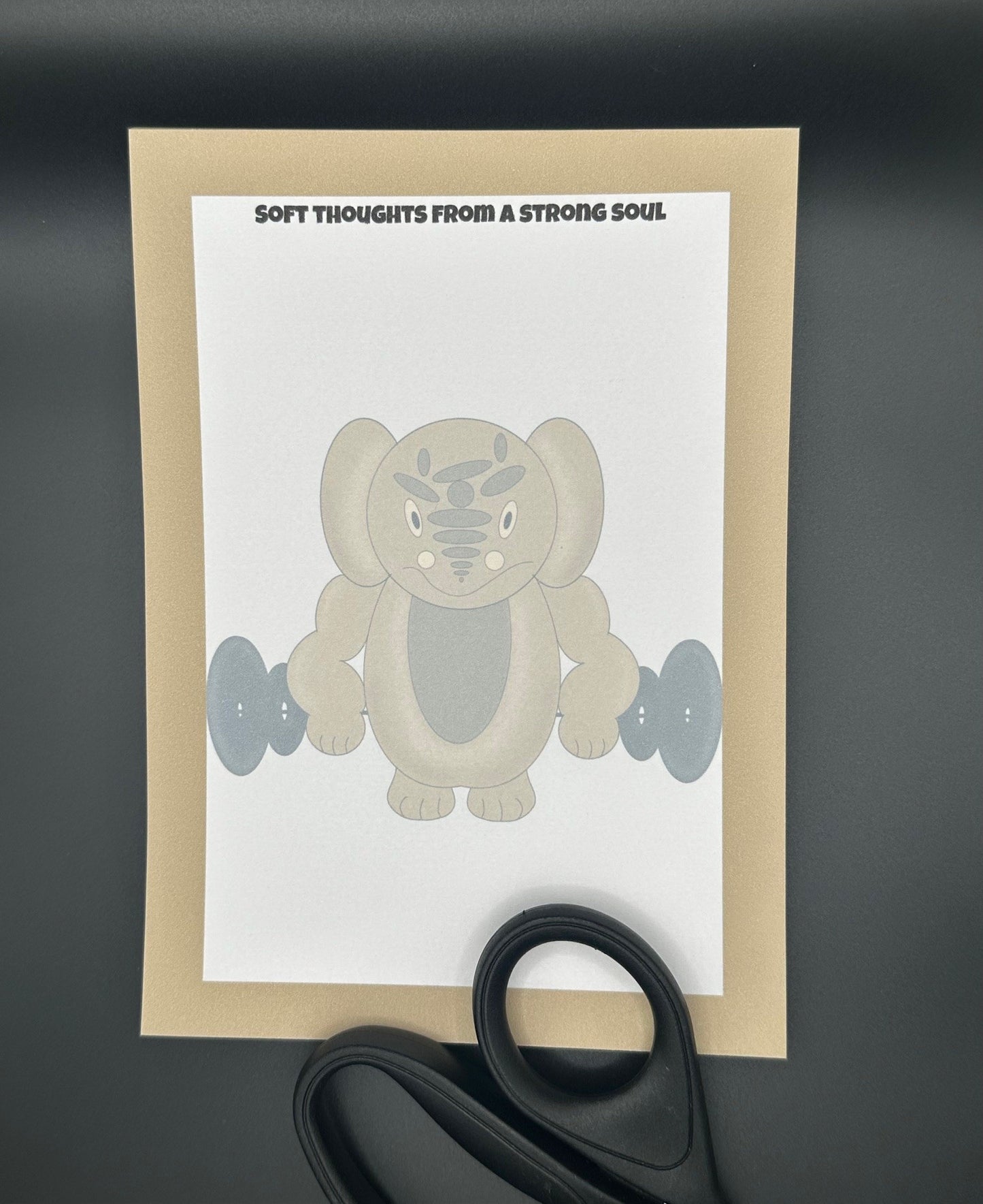 A notepad that has a brown character that is rounded shape with muscles and has black eyes and a smile. The character is holding black weights. The notepad has brown edges all around the four corners and a notepad heading stating " Soft thoughts from a strong soul" The notepad is sitting on top of a black background with scissors. 