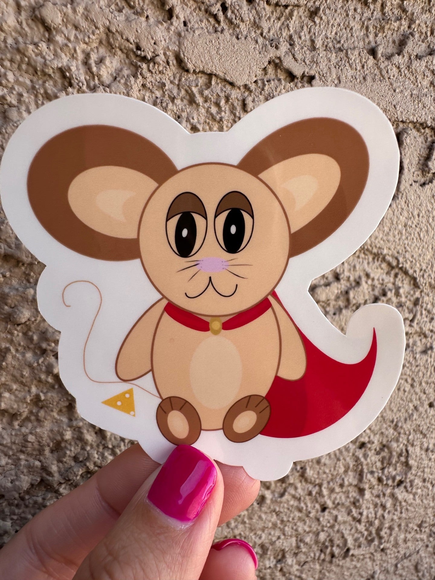 Cartoon mouse sticker with a red cape on, held by a hand with pink nail polish on a textured surface.Next to the mouse is cheese.