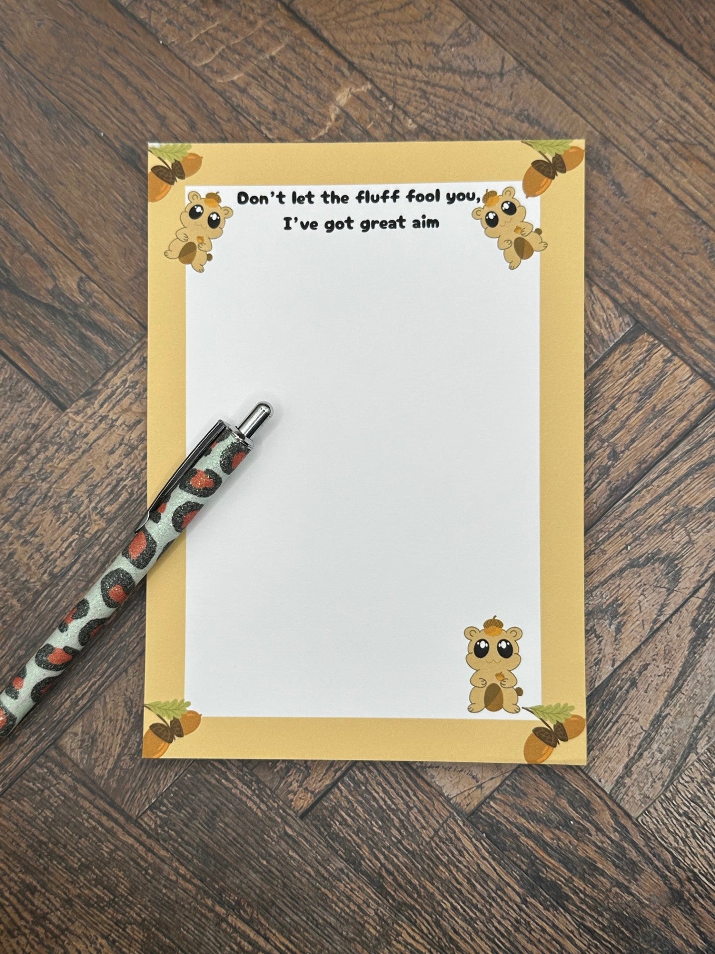 A notepad that has fictional character that is brown and has an acorn on top of the head and holding one. The character has big black eyes with white sparkles in it. The character is at the bottom of the notepad and two on top. The notepad has brown edges on all four corners of the notepad with acorns on all four corners as well. The notepad heading reading " Don't let the fluff fool you, I've got great aim". The notepad is sitting on a wooden surface with a pen.