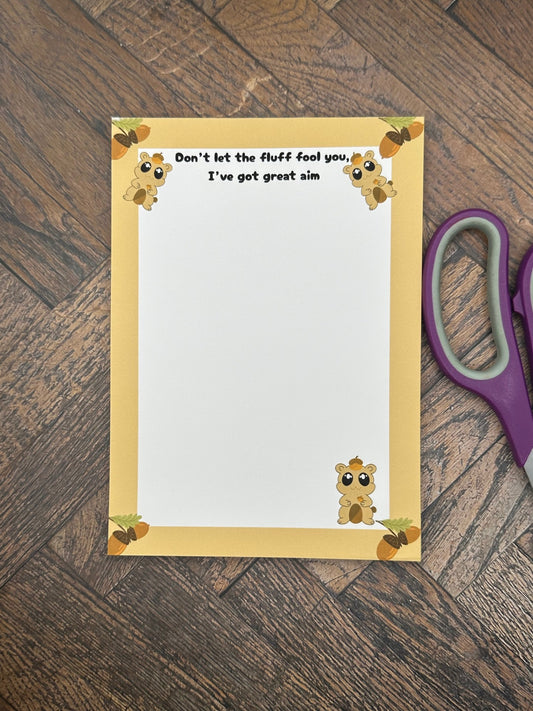 A notepad that has fictional character that is brown and has an acorn on top of the head and holding one. The character has big black eyes with white sparkles in it. The character is at the bottom of the notepad and two on top. The notepad has brown edges on all four corners of the notepad with acorns on all four corners as well. The notepad heading reading " Don't let the fluff fool you, I've got great aim". The notepad is sitting on a wooden surface with scissors. 