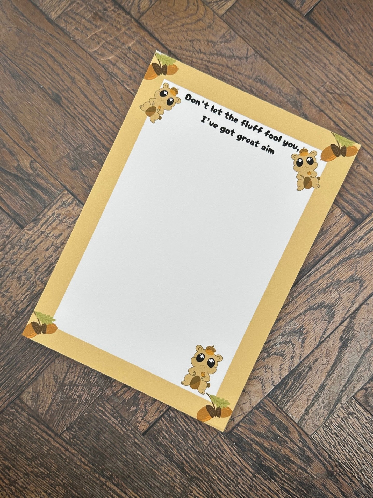 A notepad that has fictional character that is brown and has an acorn on top of the head and holding one. The character has big black eyes with white sparkles in it. The character is at the bottom of the notepad and two on top. The notepad has brown edges on all four corners of the notepad with acorns on all four corners as well. The notepad heading reading " Don't let the fluff fool you, I've got great aim". The notepad is sitting on a wooden surface. 