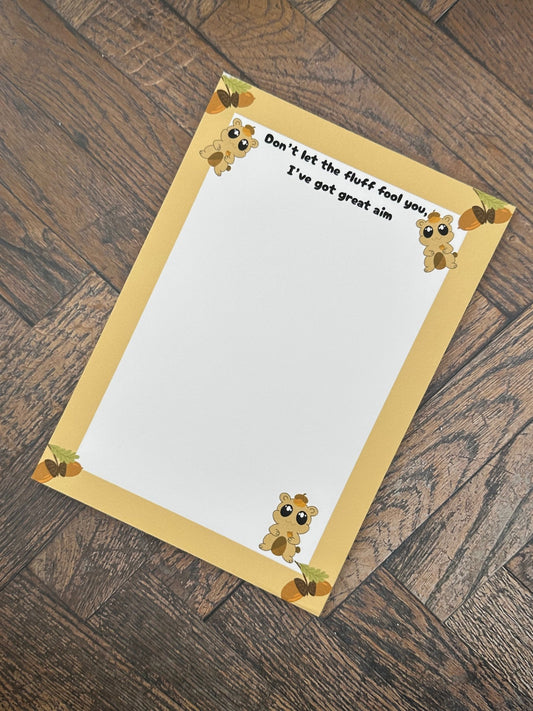A notepad that has fictional character that is brown and has an acorn on top of the head and holding one. The character has big black eyes with white sparkles in it. The character is at the bottom of the notepad and two on top. The notepad has brown edges on all four corners of the notepad with acorns on all four corners as well. The notepad heading reading " Don't let the fluff fool you, I've got great aim". The notepad is sitting on a wooden surface. 