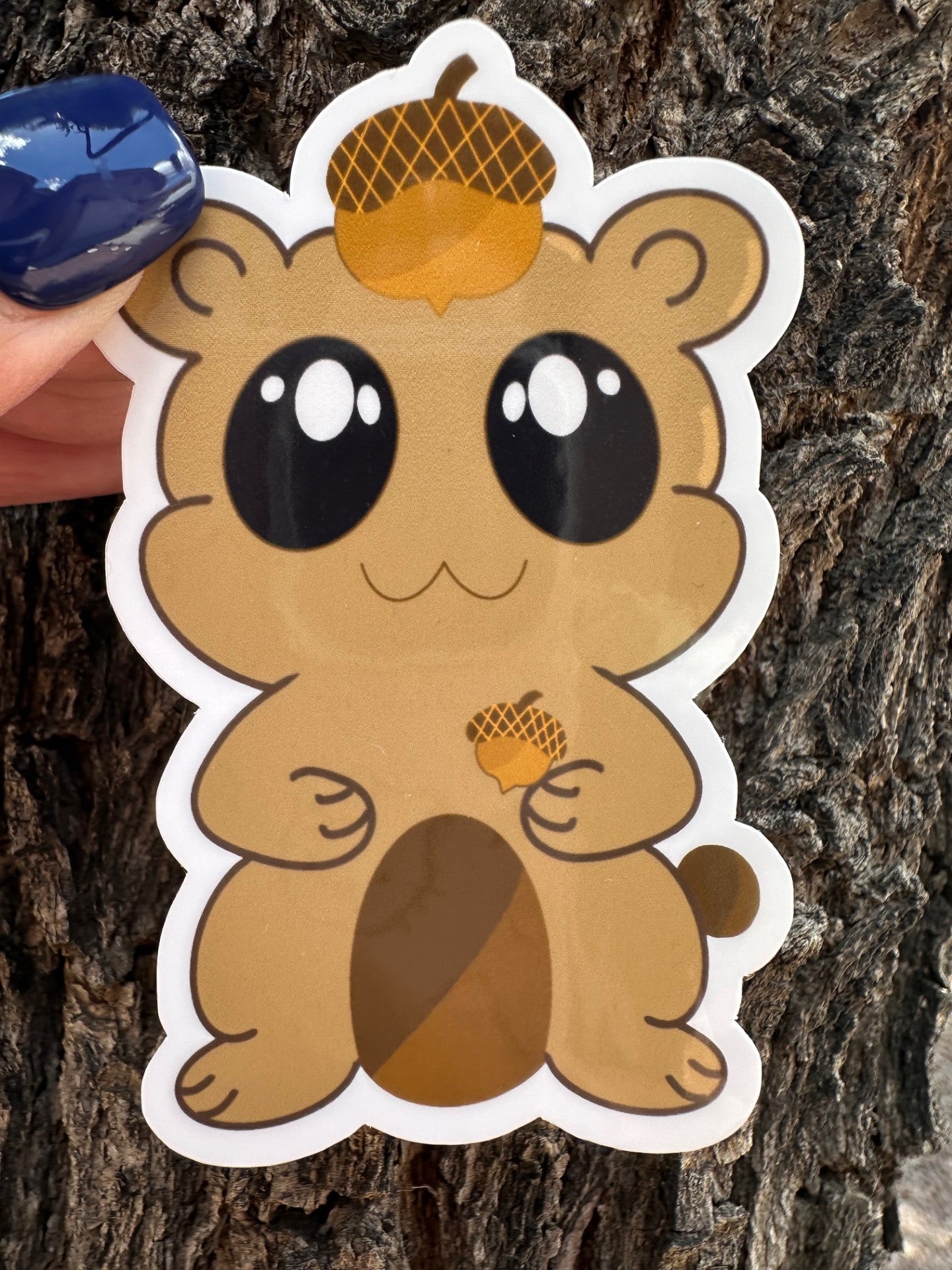 A brown fictional character glossy sticker that has big black eyes and white sparkle. The character is holding an acorn and has an acorn on top of its head. The background has a tree bark setting. 