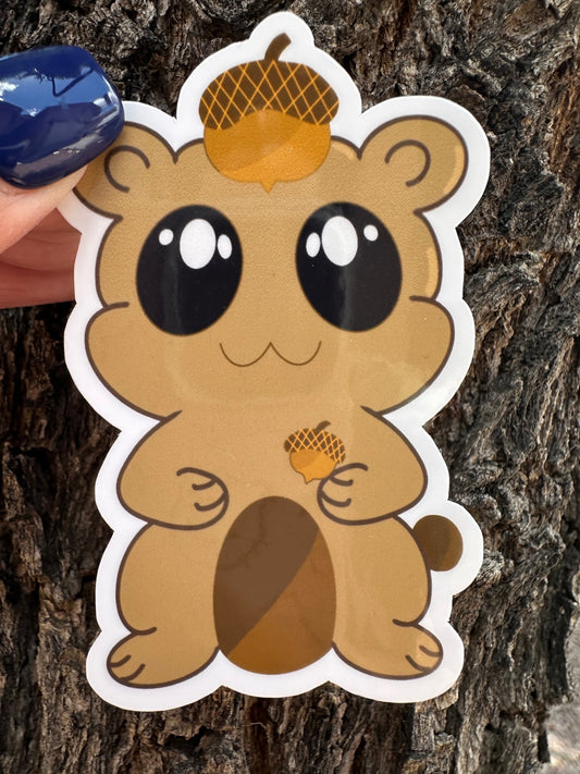 A brown fictional character glossy sticker that has big black eyes and white sparkle. The character is holding an acorn and has an acorn on top of its head. The background has a tree bark setting. 
