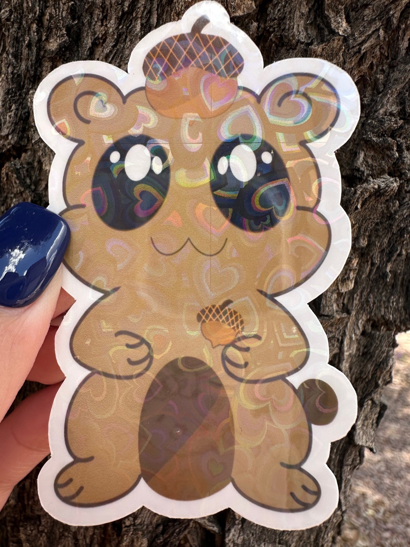 A brown fictional character heart holographic sticker that has big black eyes and white sparkle. The character is holding an acorn and has an acorn on top of its head. The background has a tree bark setting. 