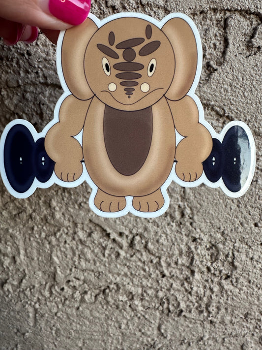 A cartoonish sticker of a brown creature that has small black eyes, and the character is holding weights, with a white border held by a hand on a textured surface. 