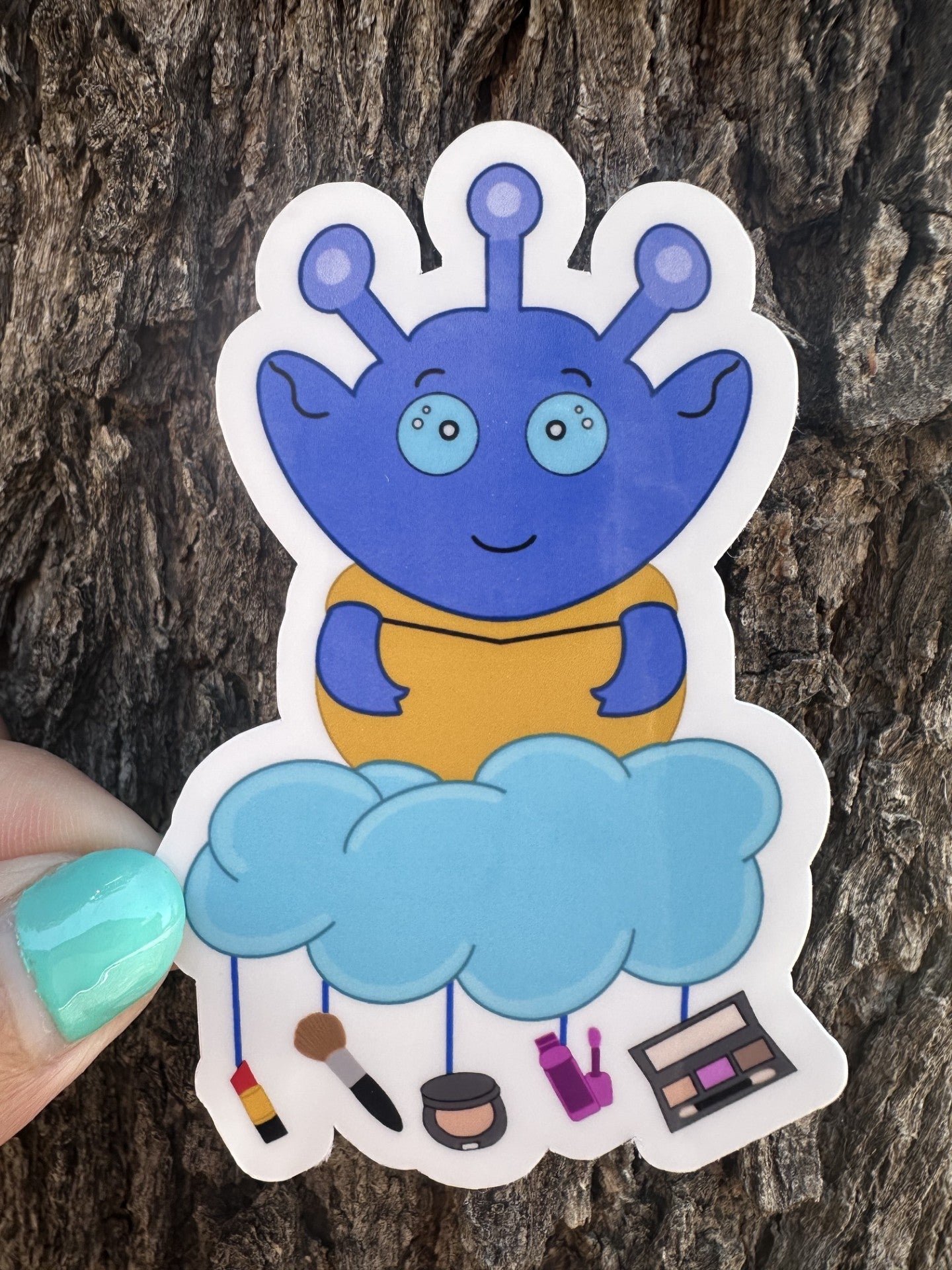 Blue alien sticker with a cloud and various makeup items hanging from the cloud. The background is a tree trunk.